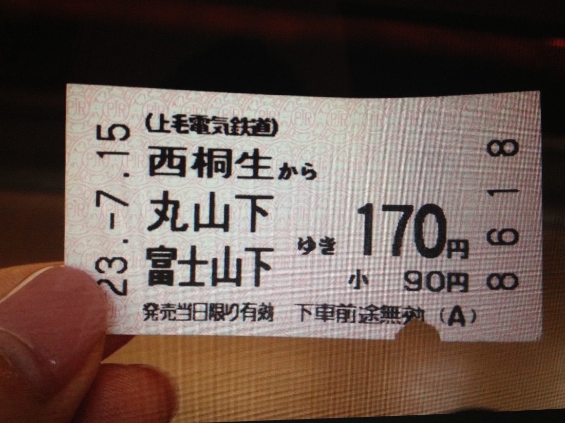 A hand holding a train ticket printed with the station names Nishi-Kiryu, Maruyamashita, and Fujiyamashita.