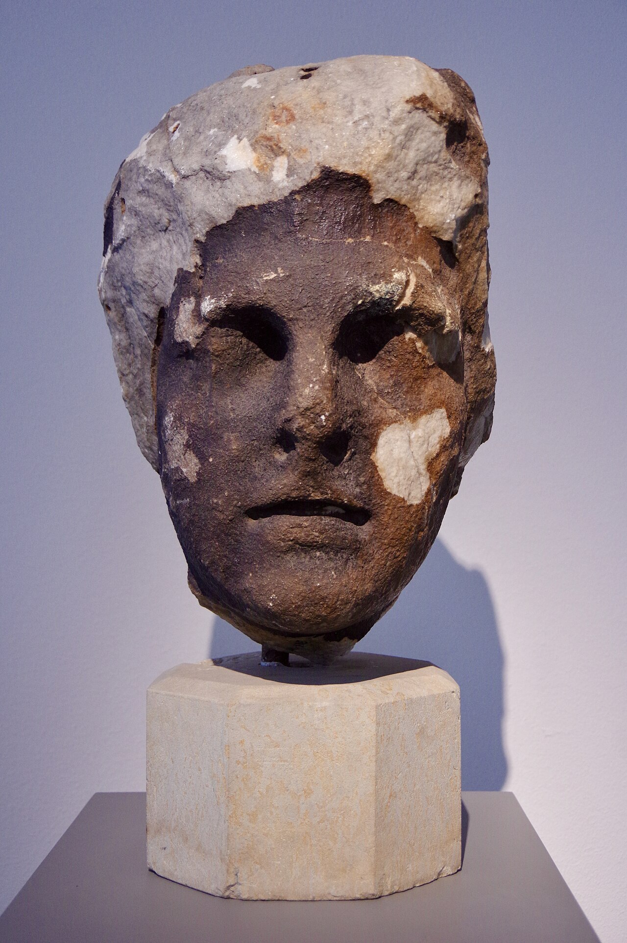Weathered marble head of Helios on a pale plinth against a lilac gallery wall