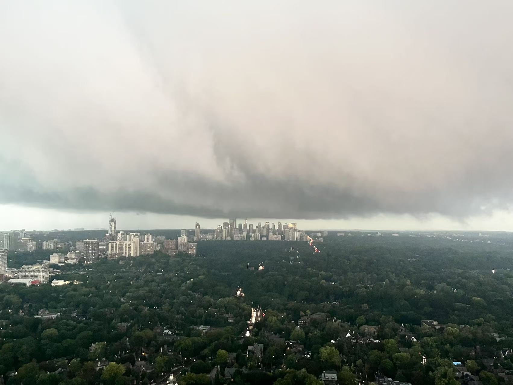 A vast shelf cloud stretched across the sky over midtown Toronto, the downtown towers small and lit beneath it, dark tree canopy in the foreground