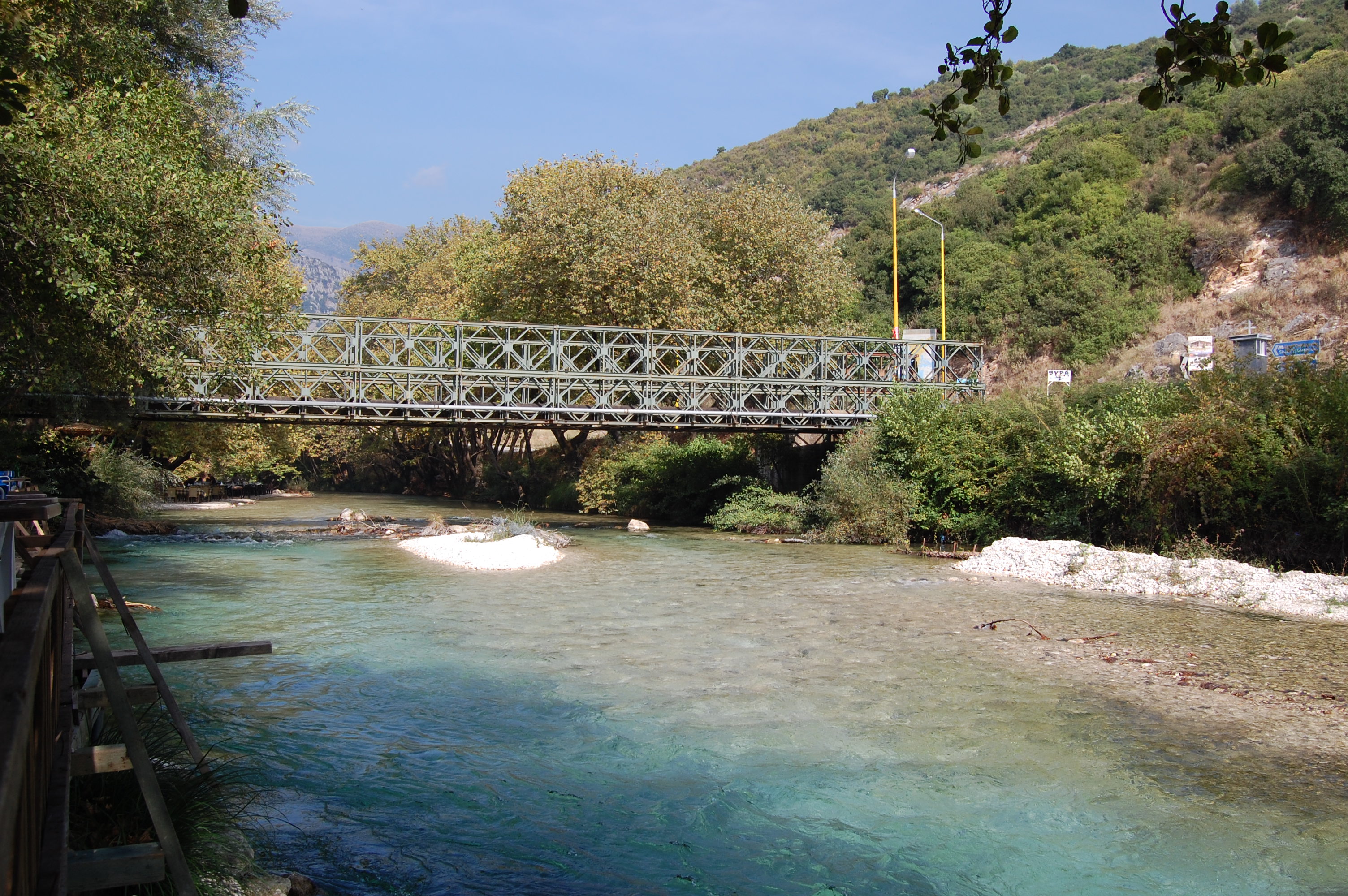 綠色鋼桁架橋橫跨清澈的河面，兩岸綠丘