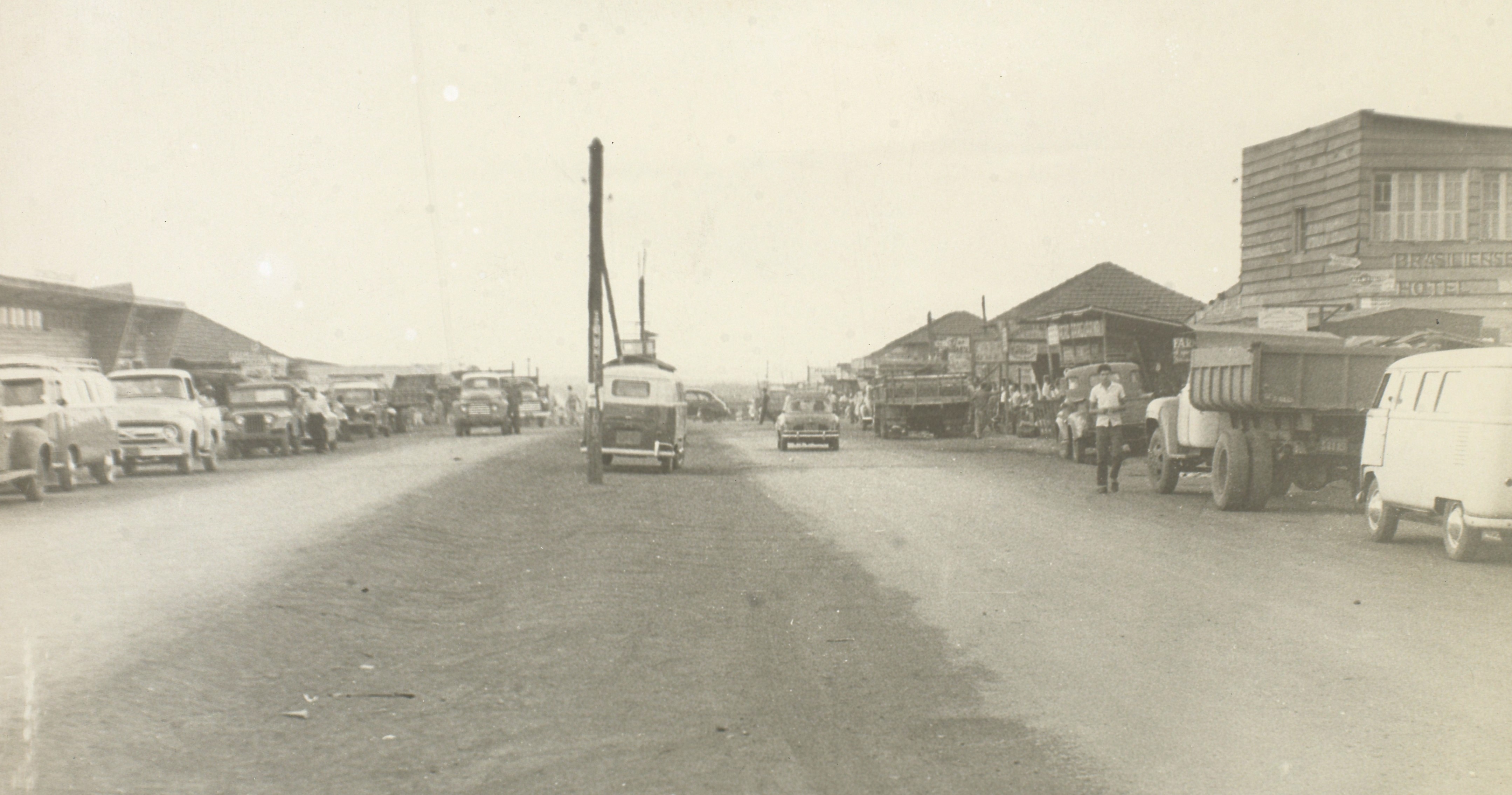 Rua larga de terra com caminhões, camionetas e pessoas diante de construções baixas de madeira; à direita um sobrado com a palavra Hotel.