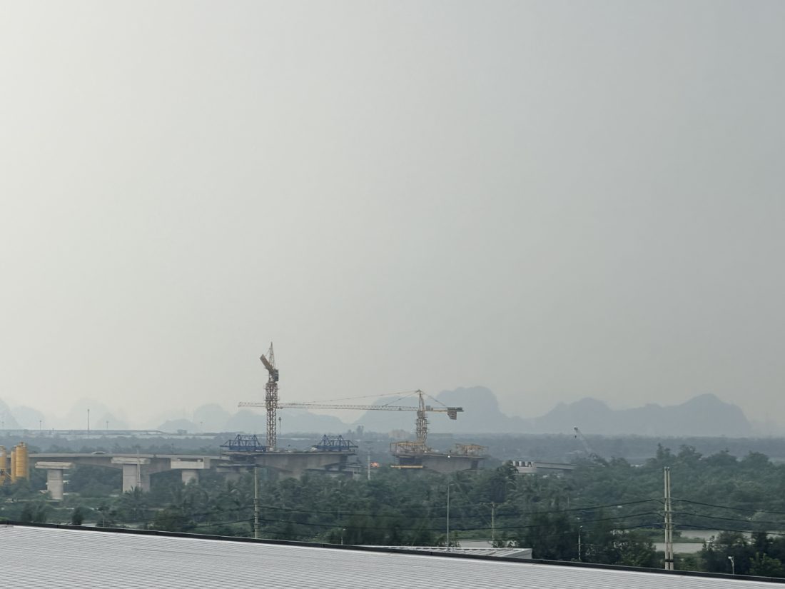 Construction cranes rising above tree canopy, limestone peaks in haze