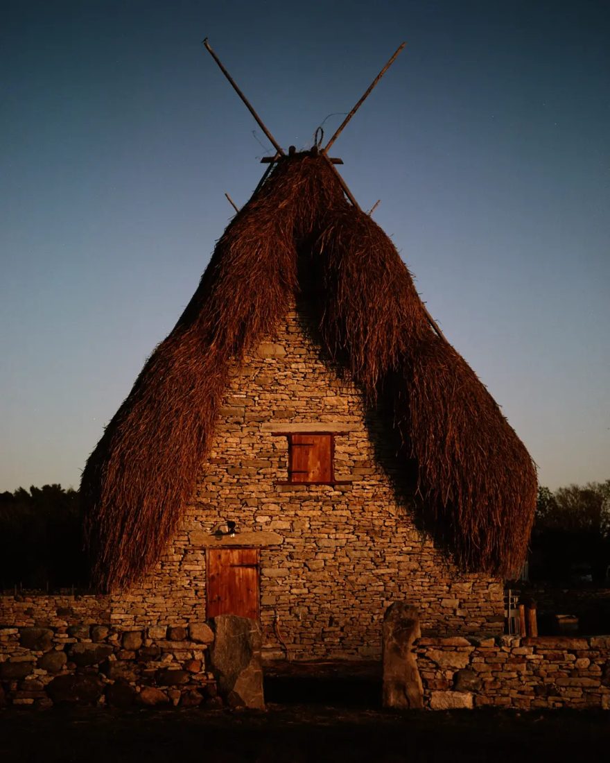 A stone house with a steep thatched roof and crossed timber poles, lit orange by the last of the evening sun under a deep blue sky.