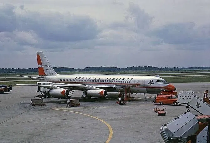 American Airlines Convair-era aircraft in polished bare metal with an orange lightning bolt stripe, parked on the apron with ground service vehicles.