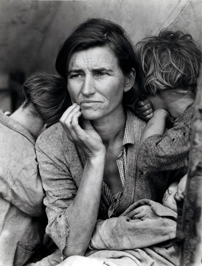 Photograph, 1936: a woman seated at the opening of a tent shelter, one hand raised to her chin, two children turned in against her shoulders with their faces away from the camera, and an infant asleep in her lap.