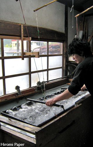 Documentary photograph: a papermaker at a wooden vat in a daylit workshop, moving a screen and frame through water clouded with plant fibre.