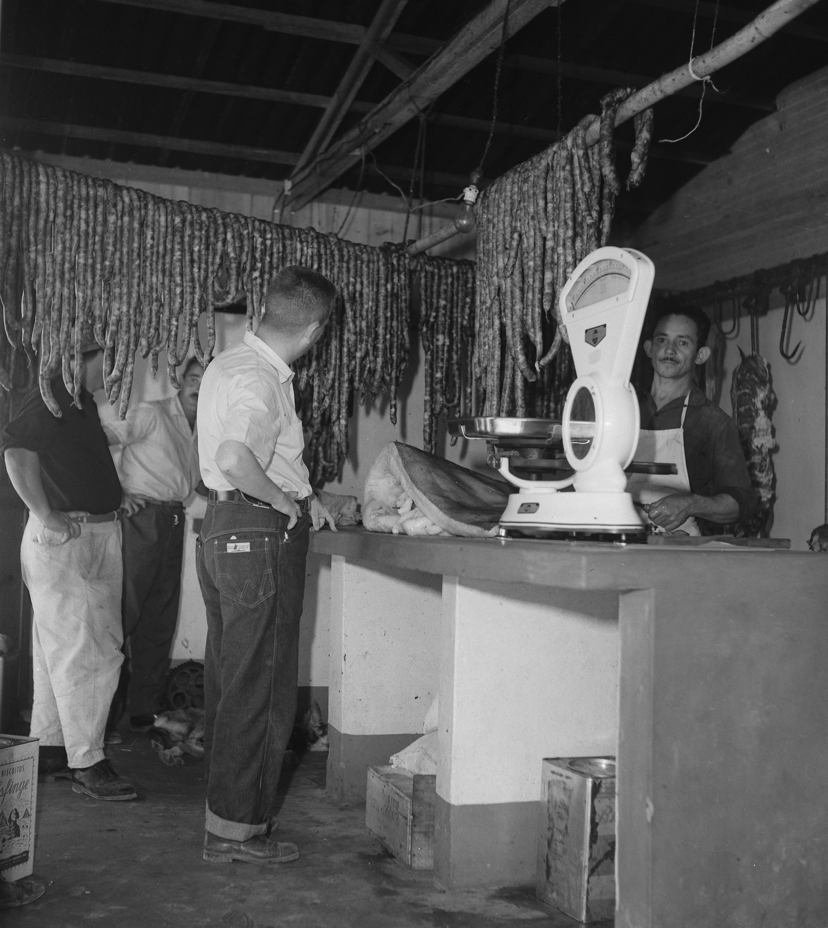 Interior de uma venda: linguiças penduradas em varas, uma balança clara sobre o balcão, um homem de avental atrás do balcão e três clientes de pé.