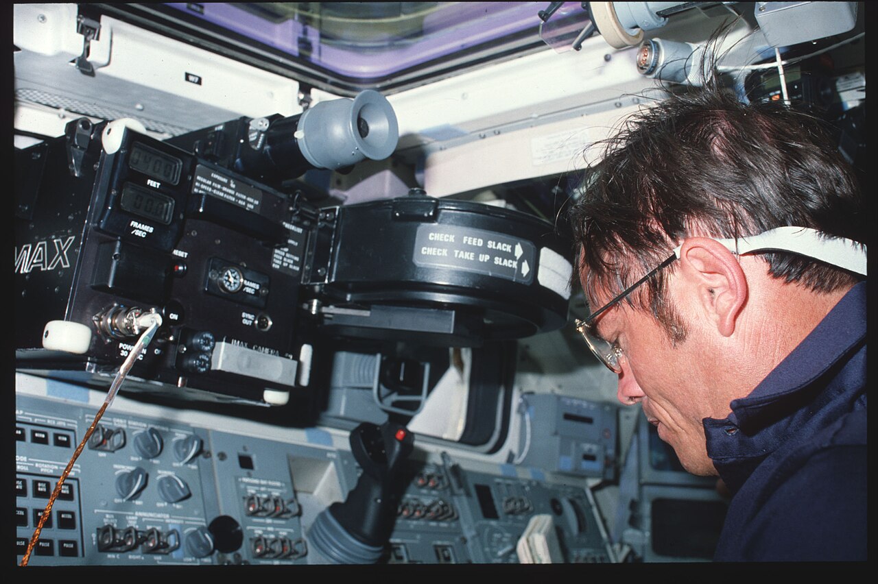 A crewman in a headset leaning over a camera on the Space Shuttle Challenger flight deck.