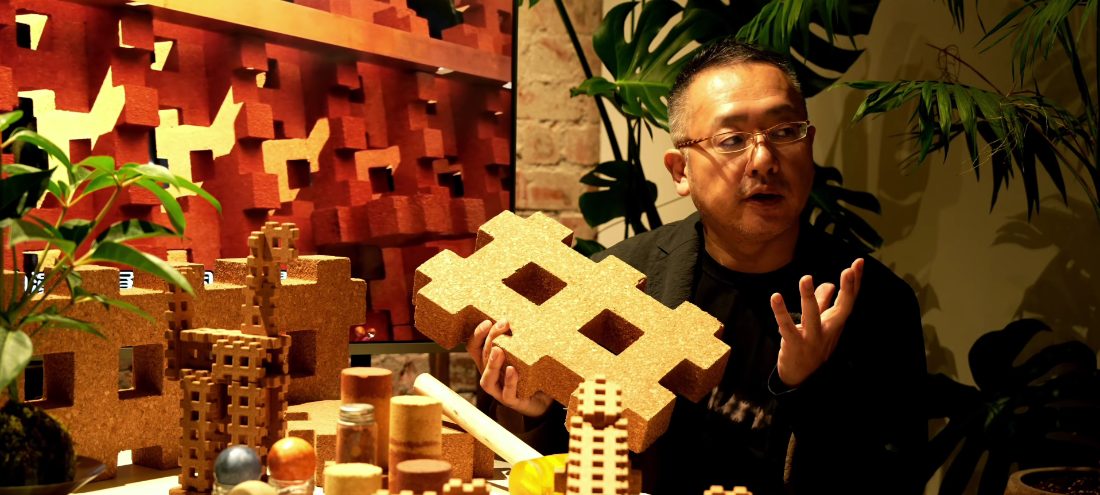 Yoshitoki Iijima holding a cork module block, speaking at an exhibition surrounded by cork structures and tropical plants