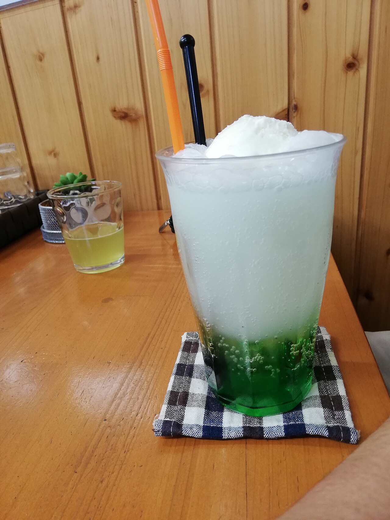 A tall glass of green melon soda topped with a scoop of vanilla ice cream, on a checked cloth coaster.
