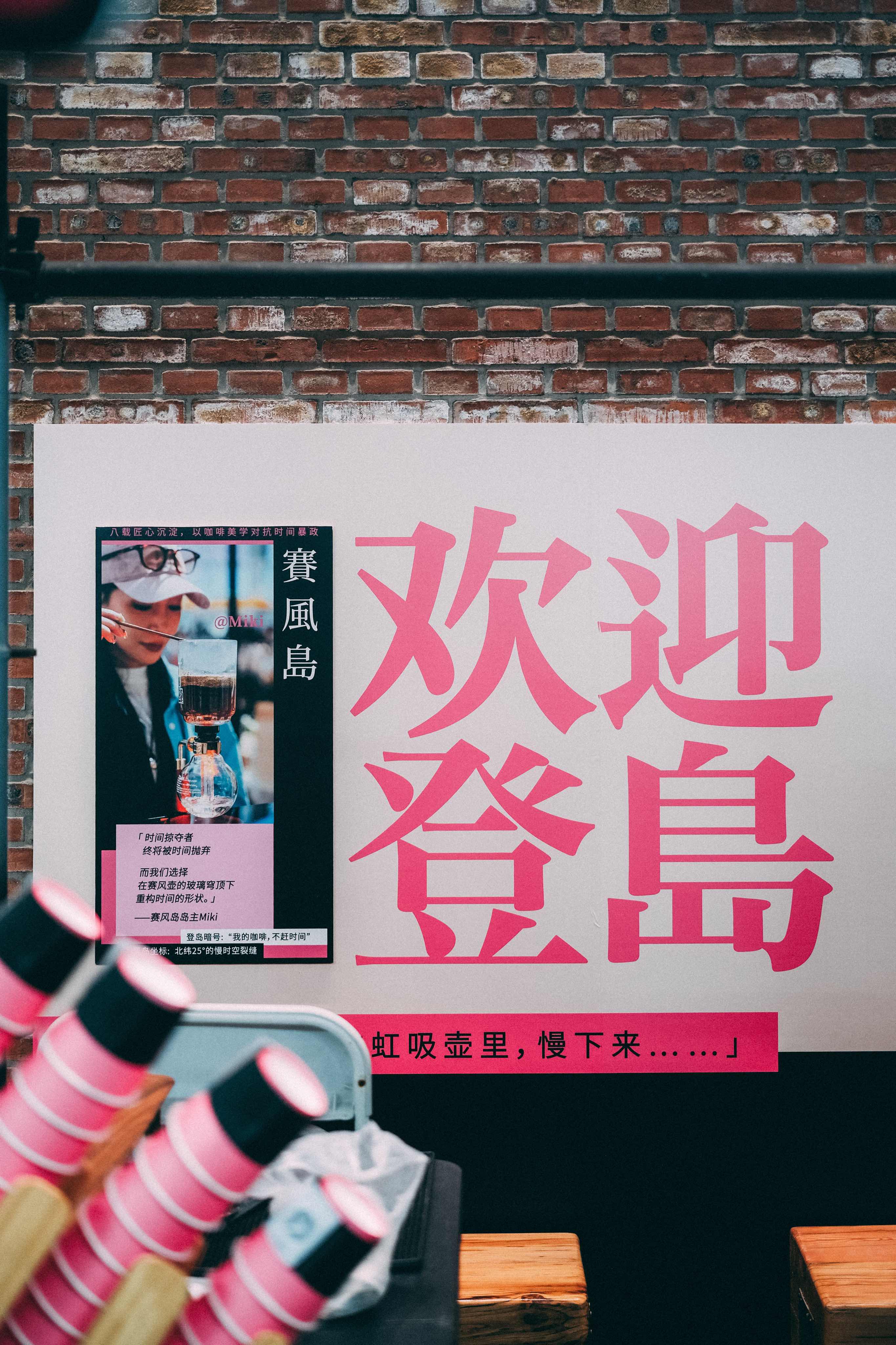 砖墙前的白色展板，粉色大字“欢迎登岛”，左侧海报上是赛风岛的品牌陈述与咖啡师照片，下方一行字写着“虹吸壶里，慢下来……”。