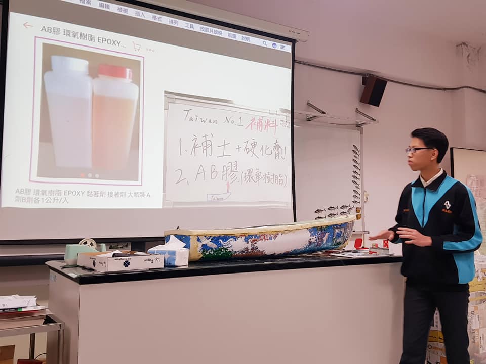 學生在教室投影 AB 膠與補土資料前，向大家說明修補一艘彩繪小船的材料。/ A student presents repair materials — filler, hardener and epoxy — beside a painted miniboat.