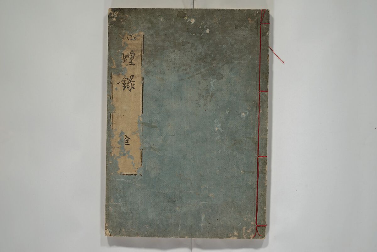 Blue-grey cover of a stitched Japanese book with a paper title label in Japanese characters and red binding thread.