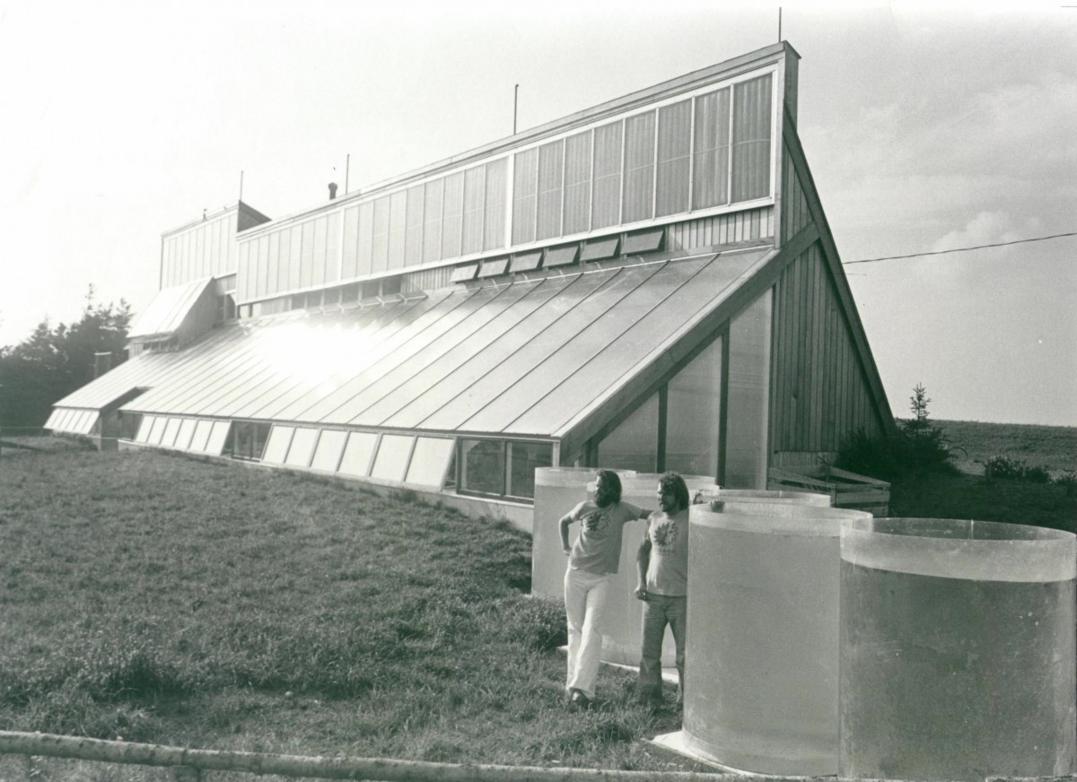 The PEI Ark seen from outside: a long, low structure with a south-facing glazed roof slope, two men standing beside cylindrical aquaculture tanks at the southeast corner, autumn 1976.