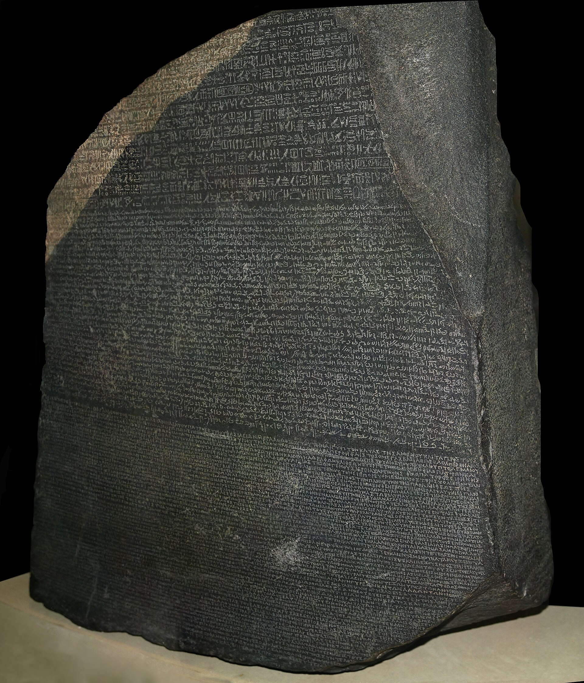 The Rosetta Stone: a broken dark granodiorite slab densely inscribed with three bands of text, the upper band in hieroglyphs, the middle in cursive Demotic, the lower in Greek capitals.