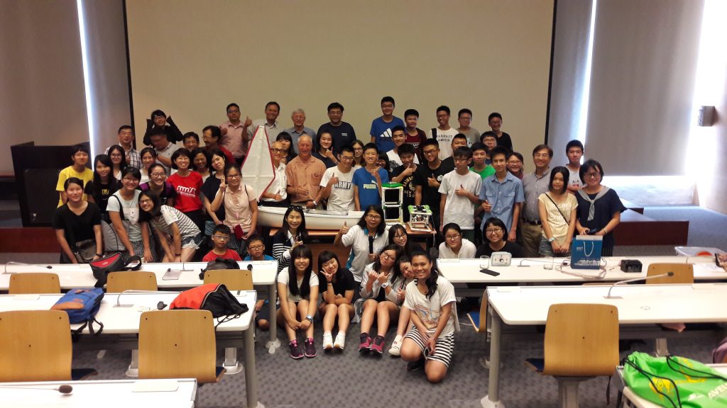 教室裡數十位學生與師長合照，前方擺著一艘白色迷你帆船 / Dozens of students and teachers in a lecture hall posing with a white mini sailboat