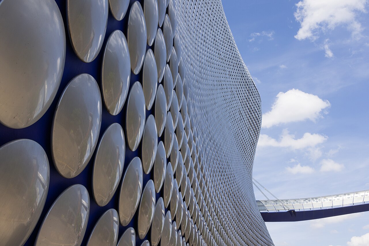 Curving building facade covered in thousands of shining aluminium discs, with a tubular walkway bridging into it, against a blue sky