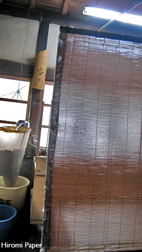 Documentary photograph: a bamboo screen hanging in the workshop with a newly formed sheet on it, beside buckets and a window.