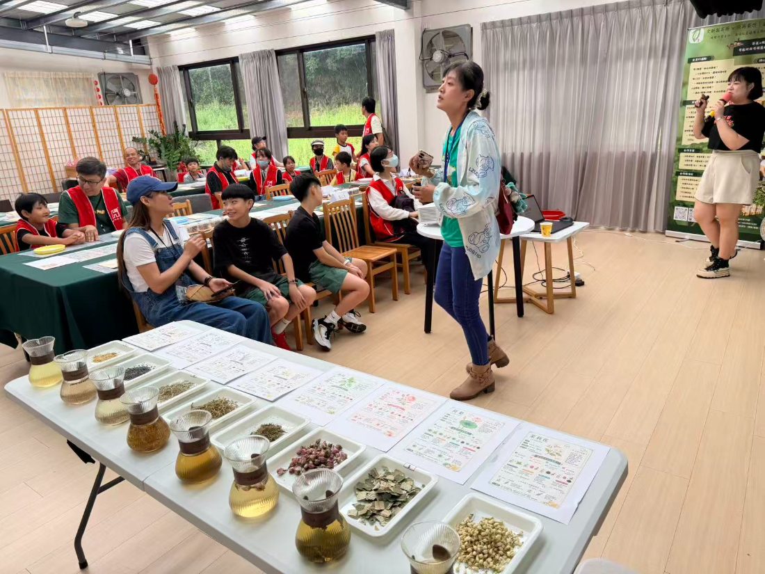 A teacher explaining a long table of dried flowers, herbs and glass pots of infusions to seated families