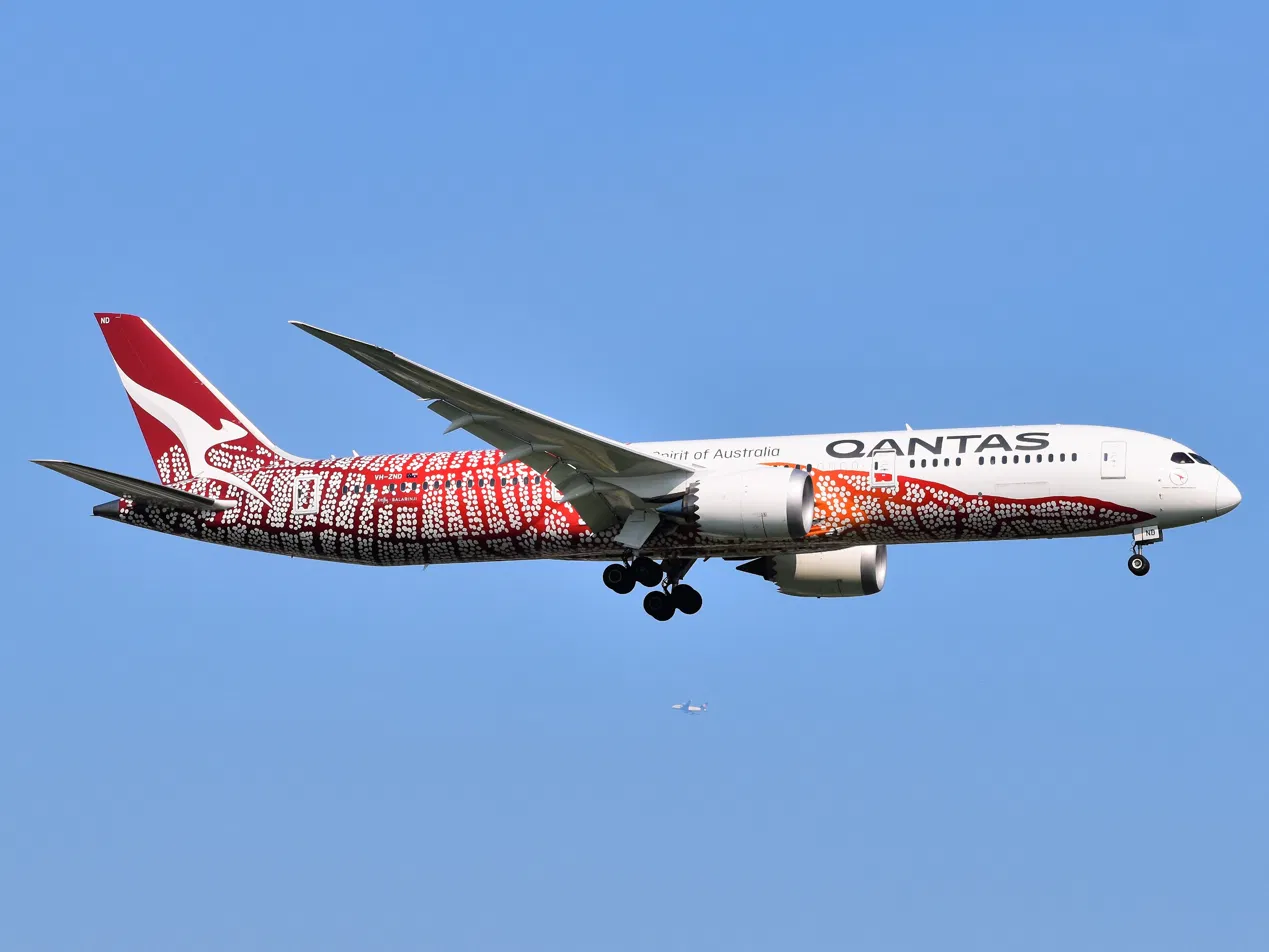 Qantas Boeing 787-9 in the Yam Dreaming livery, red and white ochre dot-work along the fuselage and tail, flying against a clear blue sky.